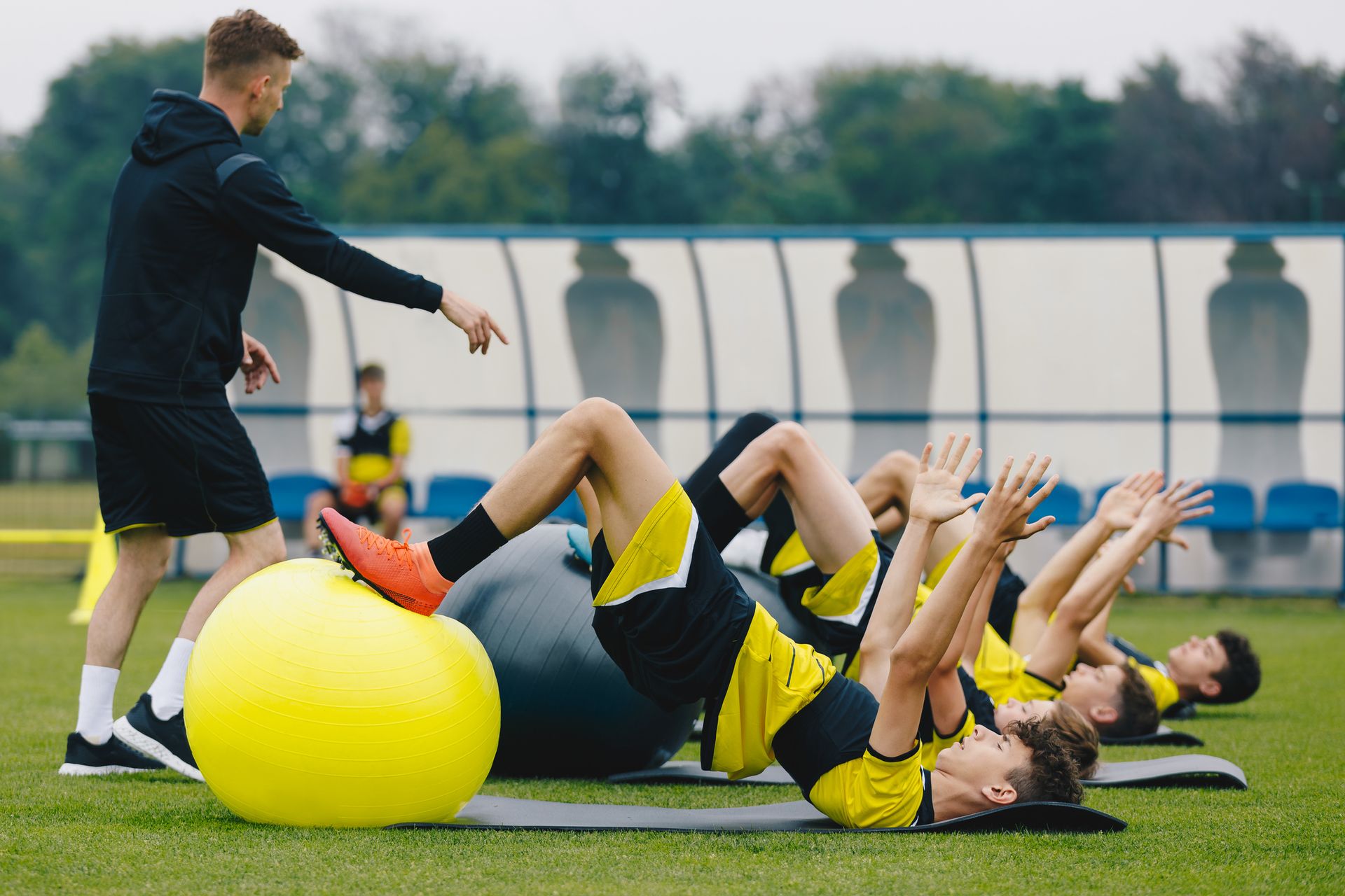 Soccer players do crunches with exercise balls on a field while a coach directs them.