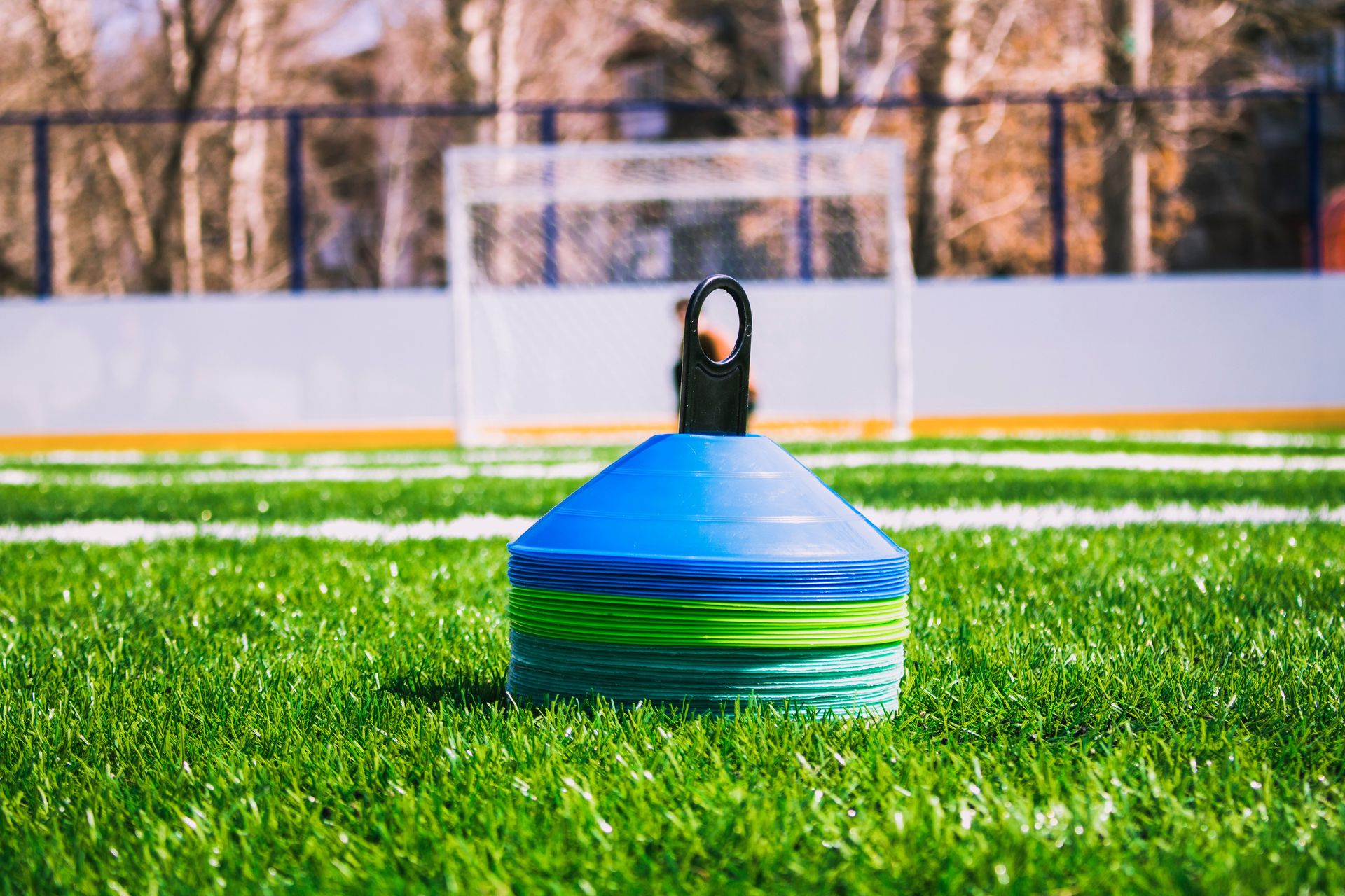 A stack of blue and green training cones sits on artificial turf in front of a goal.