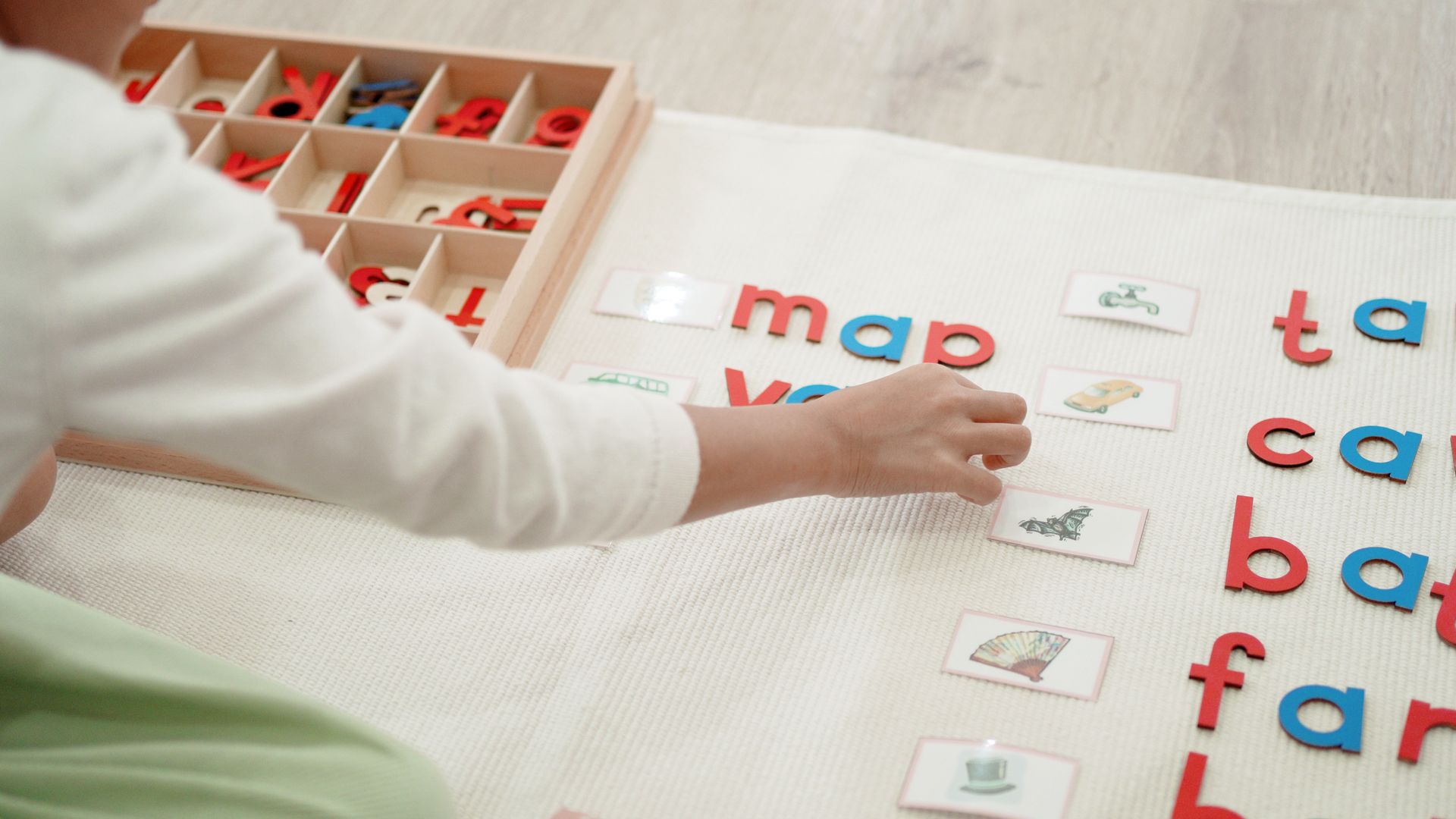A child spells short words with red and blue wooden letters next to picture cards on a white mat.