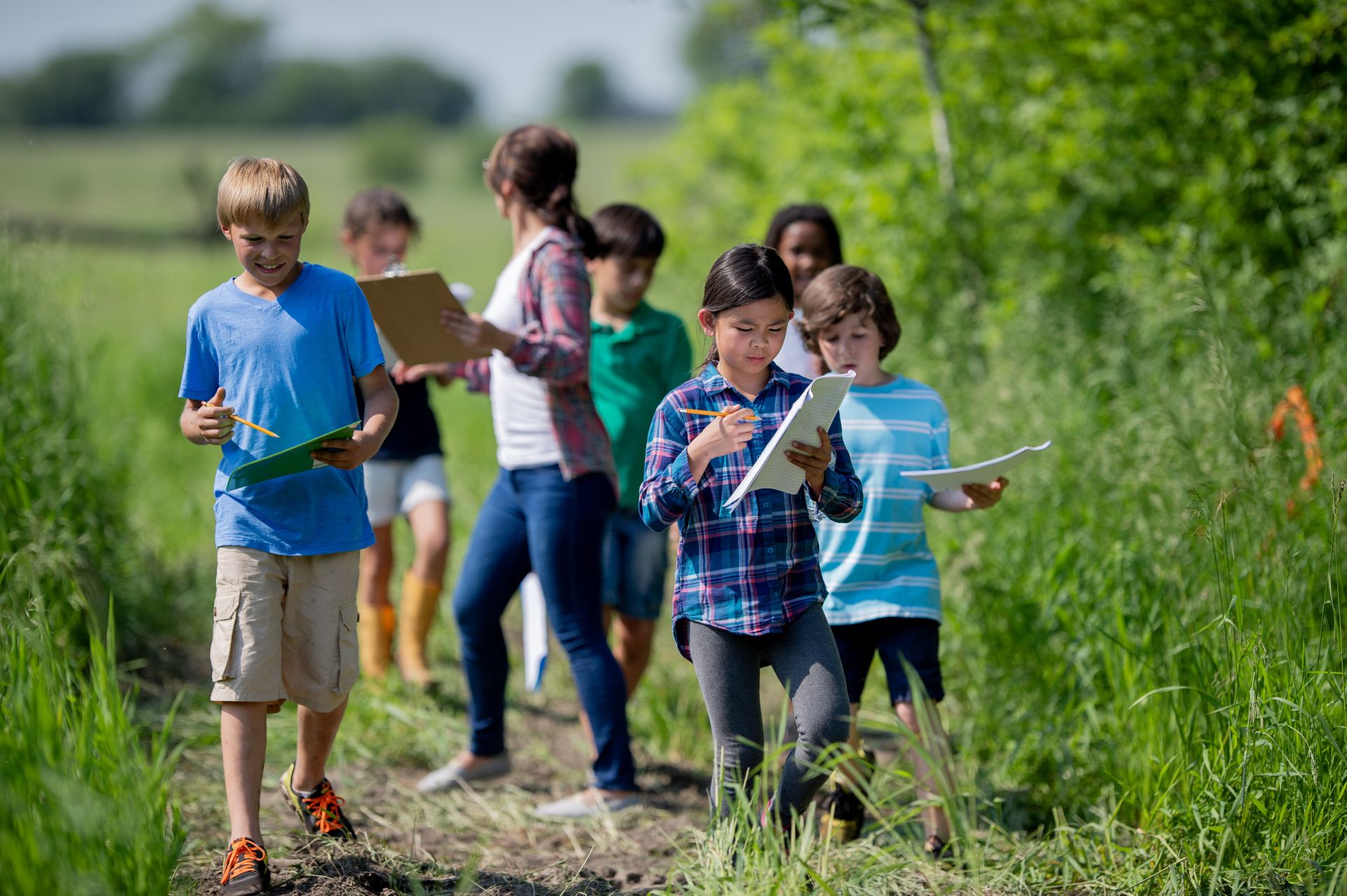 A teacher and a group of children walk along a grassy path, taking notes on clipboards and paper.