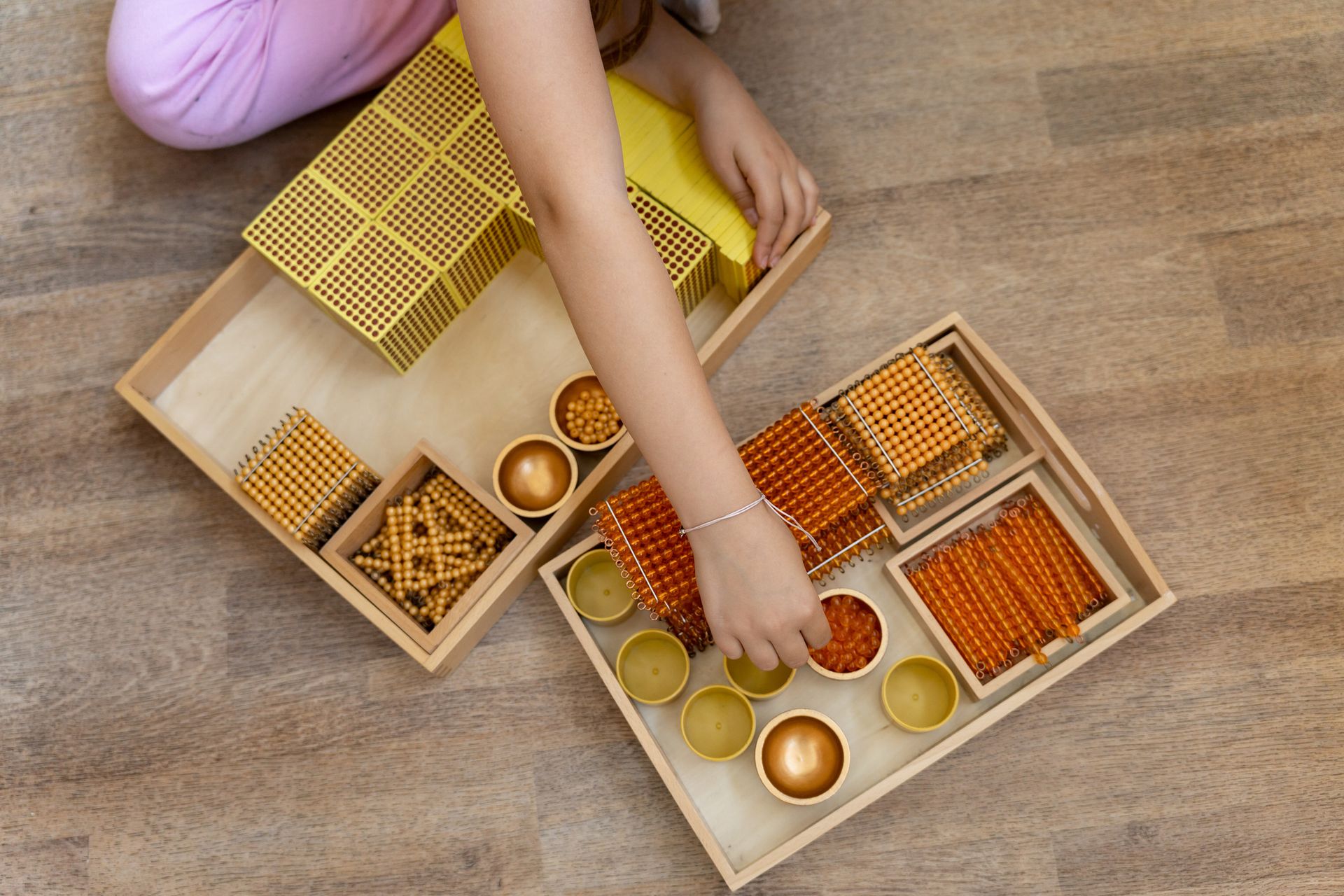 A child arranges golden bead squares and bars from a wooden tray on the floor.