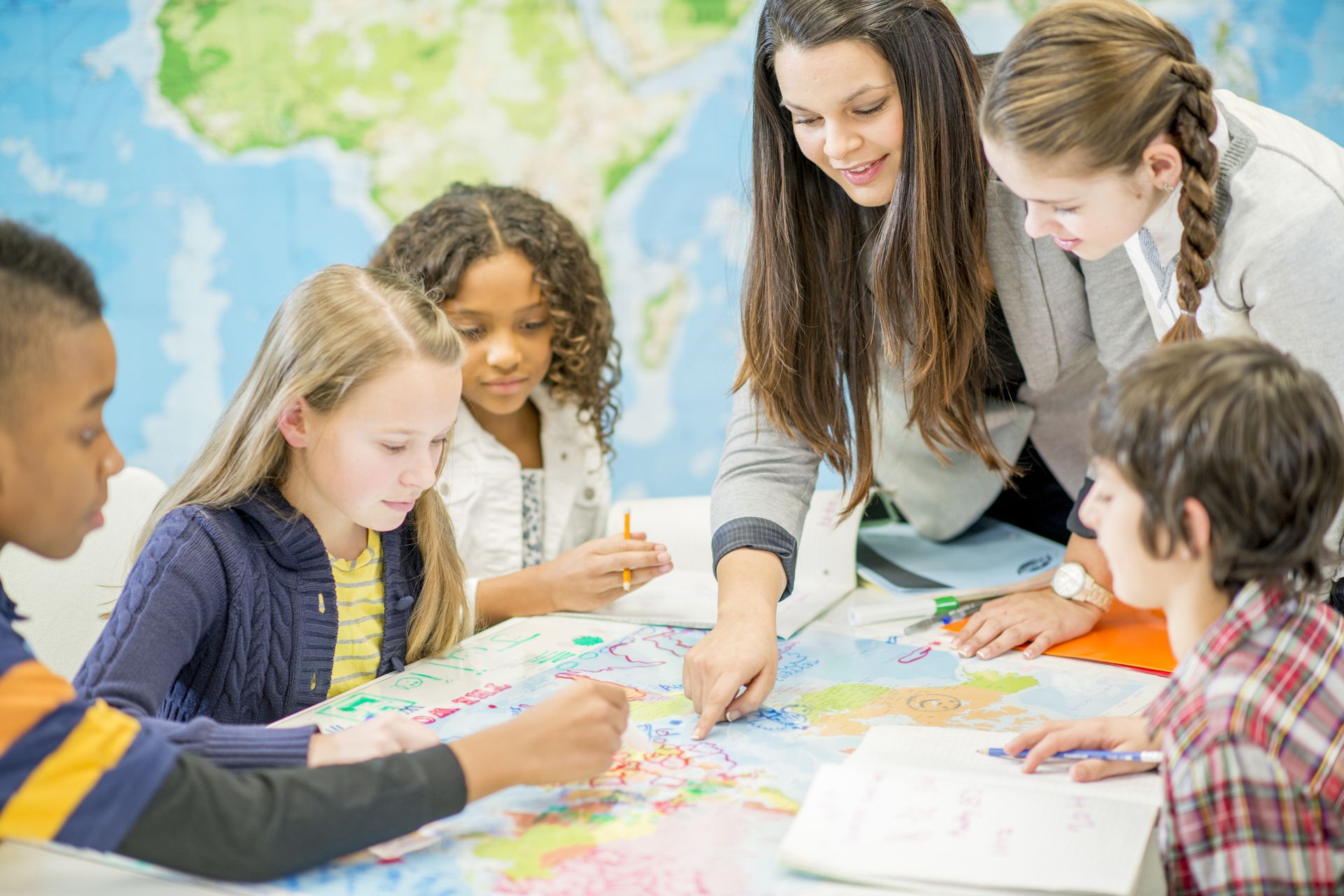 An adult points to a large world map on a table as a group of children leans in.