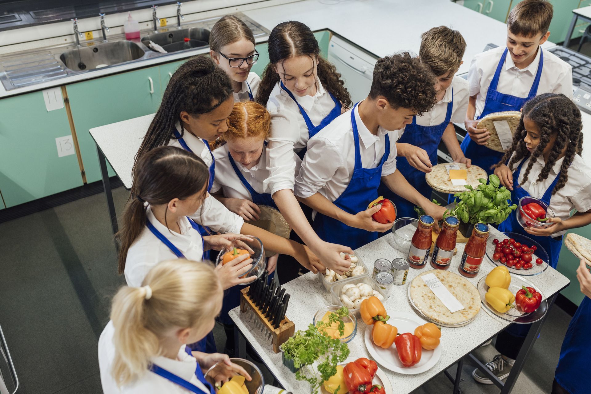 Teenagers in aprons prepare vegetables and ingredients together in a teaching kitchen.