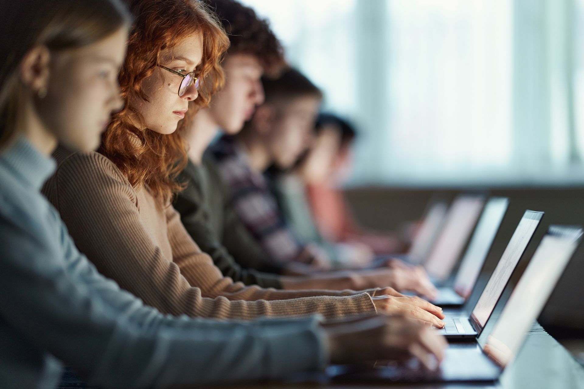A row of teenagers works on laptops at a long desk.