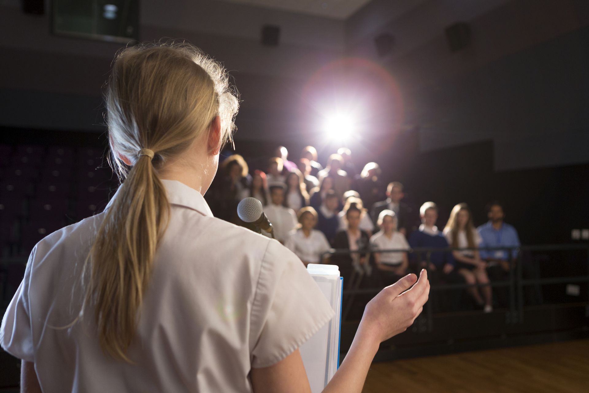 A teenager speaks from a stage under bright lights to a seated audience.