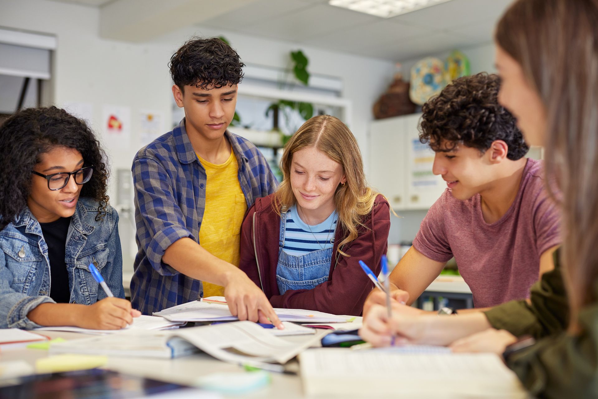 Teenagers share notes and textbooks around a classroom table.