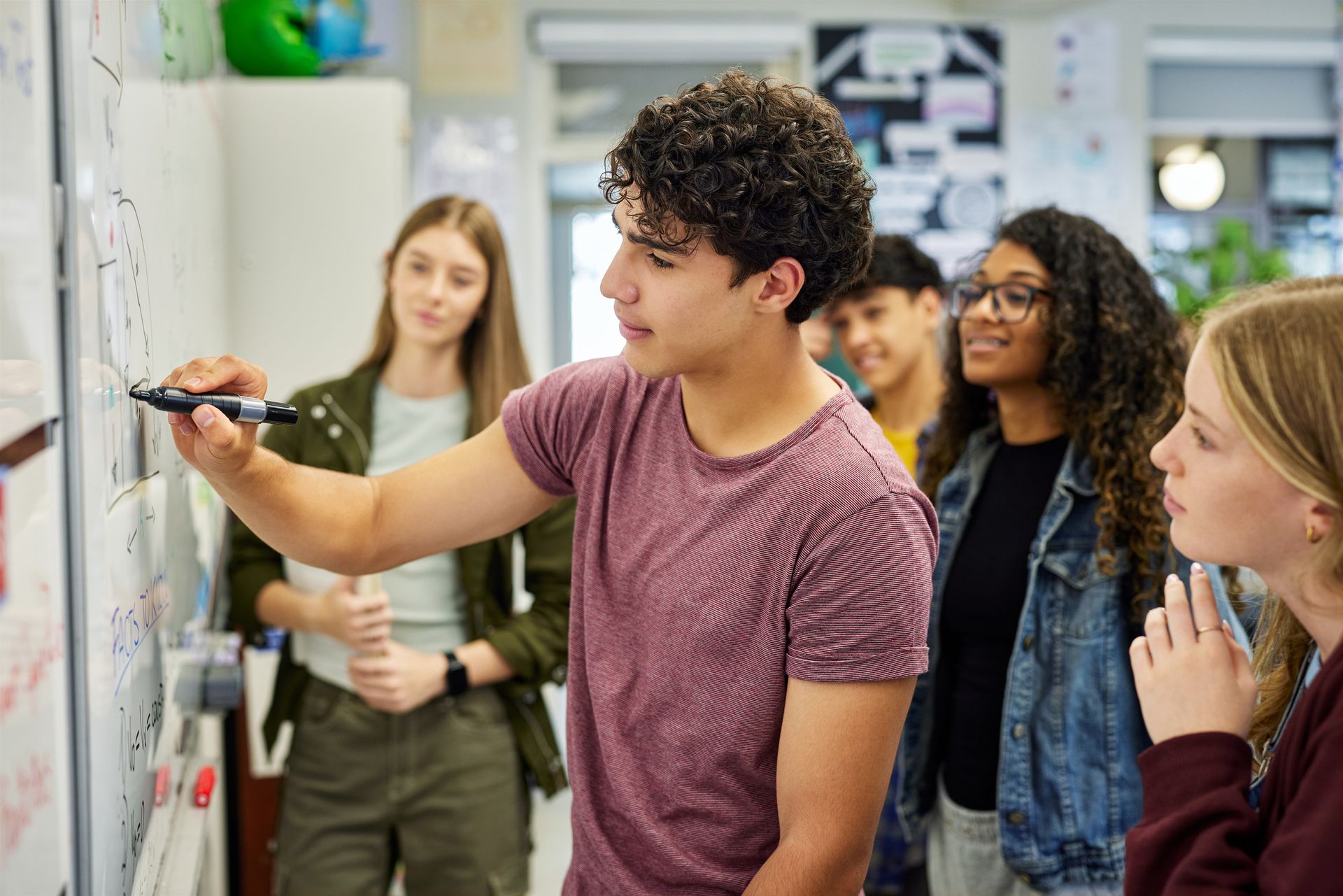 A teenager writes on a whiteboard while classmates watch and listen.